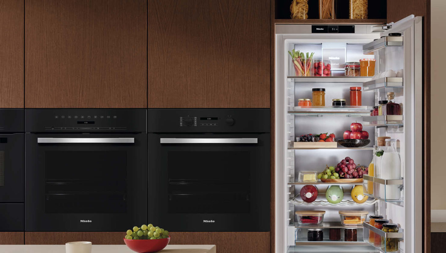 Ein offener Kühlschrank mit Gemüse, Obst, Marmeladen und Getränken von Miele in einer warmen, dunklen Holzküche.