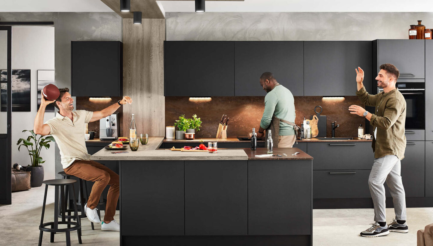 Drei junge Männer gemeinsam in einer schwarzen U-Küche: Zwei spielen mit einem Football während ein dritter kocht.