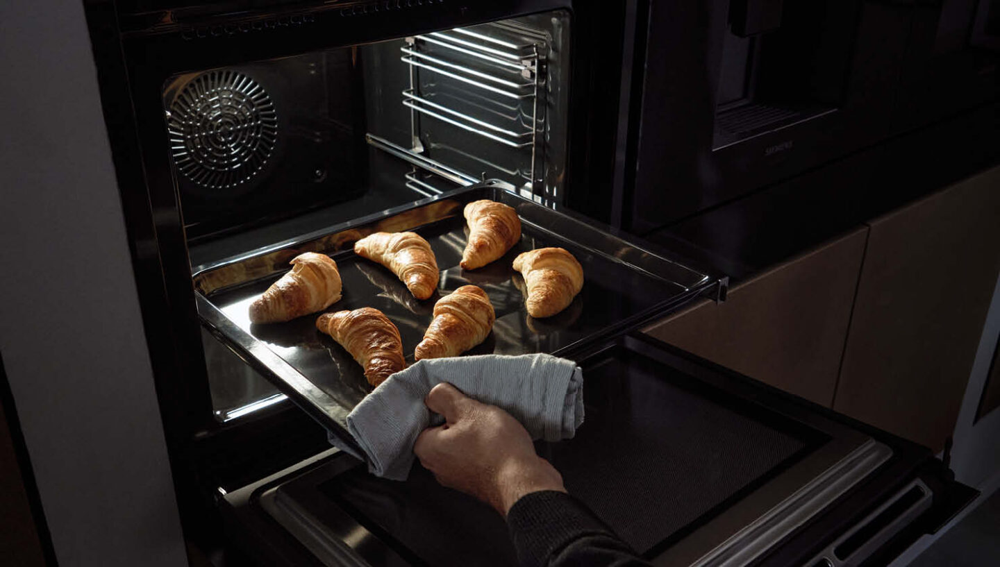 Eine Hand, die frisch gebackene Croissants aus einem Ofen von Siemens holen.