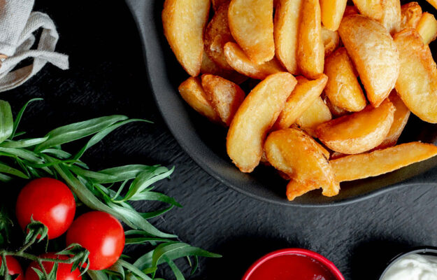 Kartoffelspalten auf einem schwarzen Teller auf einer Schieferplatte mit Ketchup und Mayonaise und Tomaten.