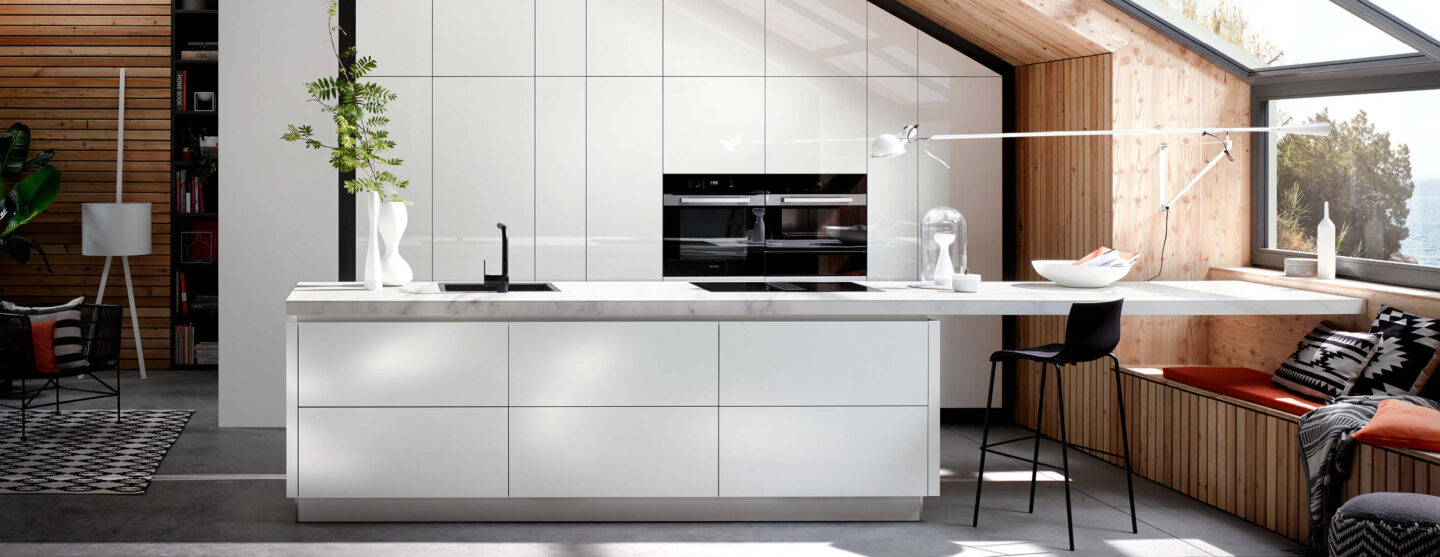 Minimalistische Designküche mit weißen Hochglanzfronten, großen Fenstern und gemütlicher Sitznische.