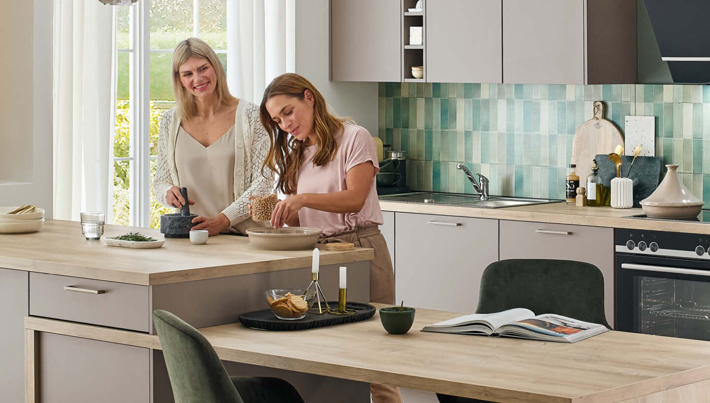 Zwei Frauen kochen gemeinsam in einer modernen, grauen Küche mit Holzarbeitsplatte. – Küche als Wohlfühlort