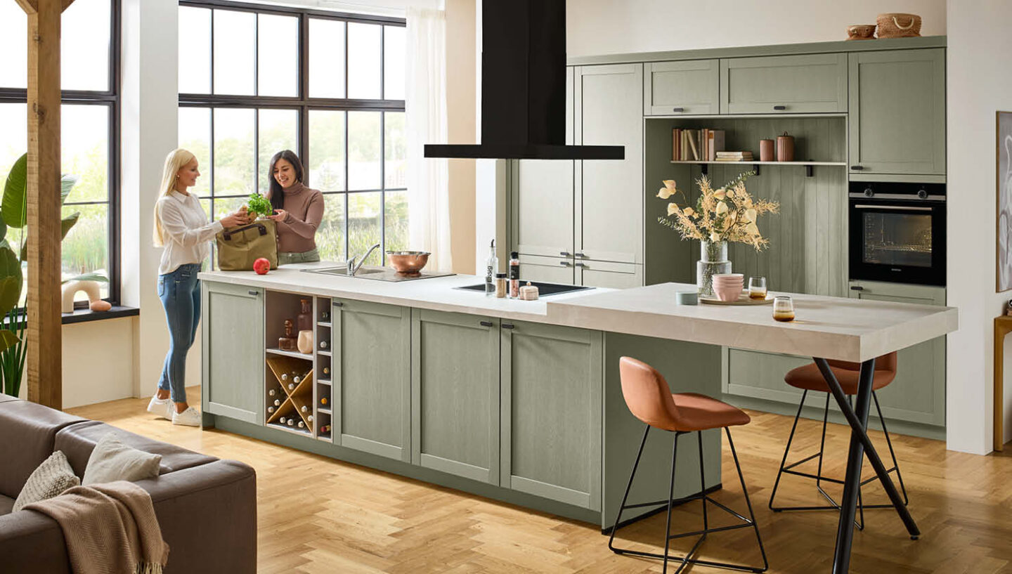 Hellgrüne Landhausküche mit weißer Steinarbeitsplatte. Zwei Frauen packen darin Einkäufe aus.