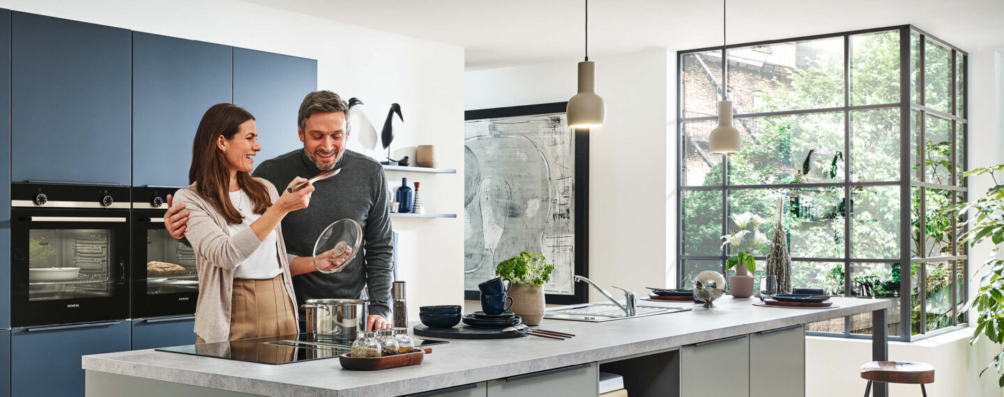Die Küche als Wohlfühlort: Eine Frau und ein Mann kochen gemeinsam in einer modernen, blauen Küche.