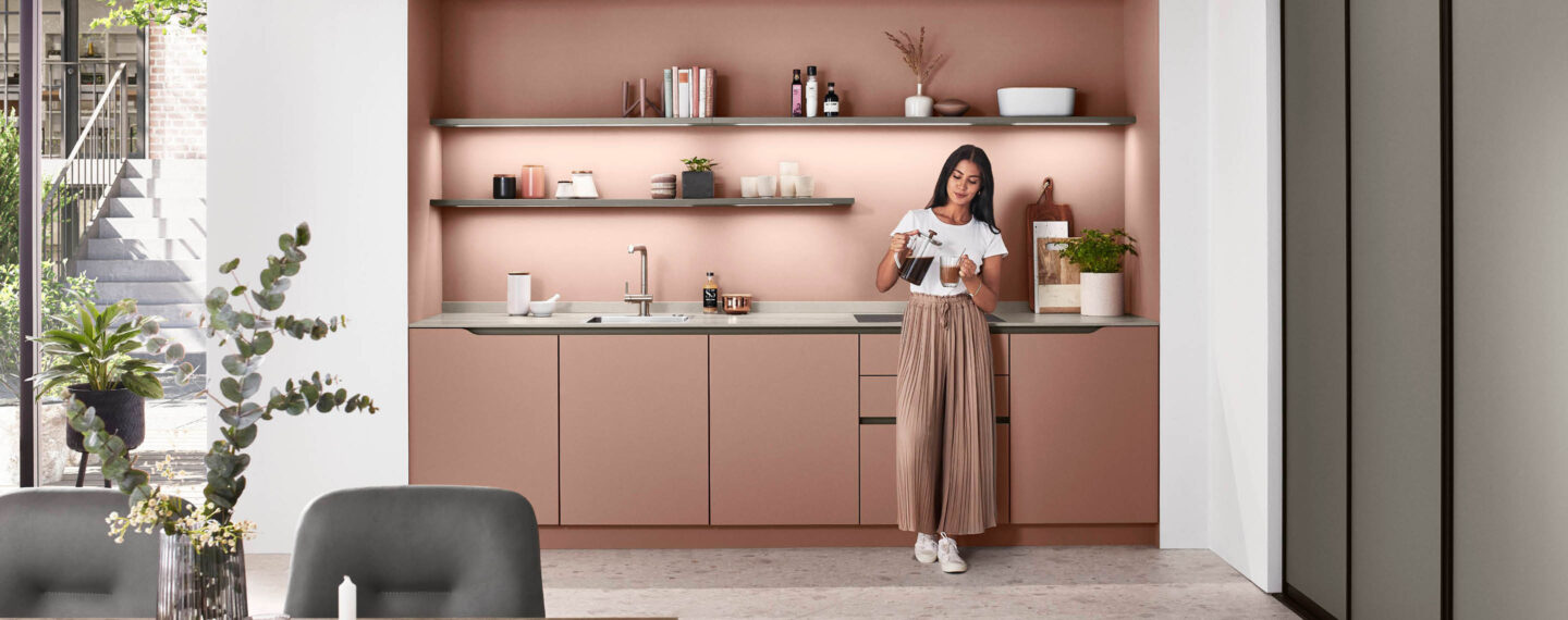 Frau steht in rosa Küchenzeile von nobilia und gießt sich einen Kaffee aus der Glaskanne ein