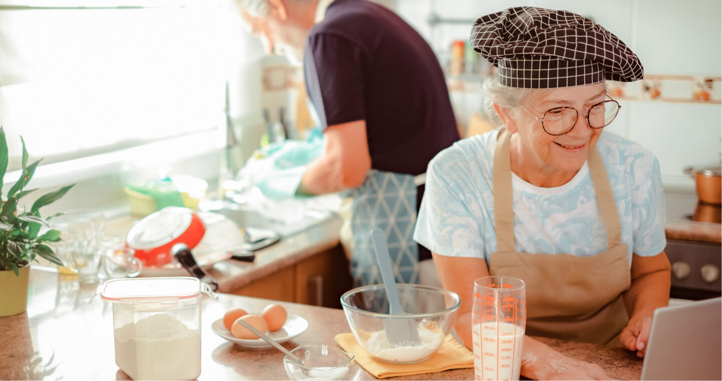 Ältere Frau beim Backen in der Küche mit Eiern, Mehl und einer Rührschüssel auf der Arbeitsfläche